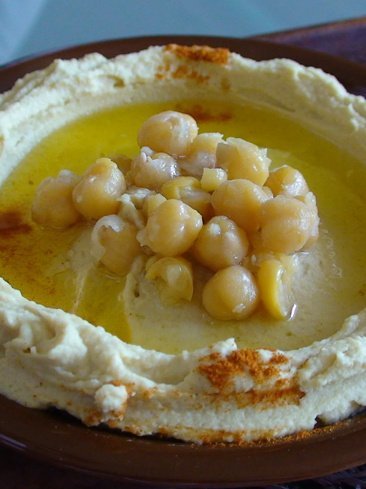 Hummus: Lebanese-style hummus. Hummus is in a brown ceramic bowl, with an indentation at the center containing olive oil and chickpeas. Hummus is sprinkled at the edges with paprika. Photo by Mr Hassan